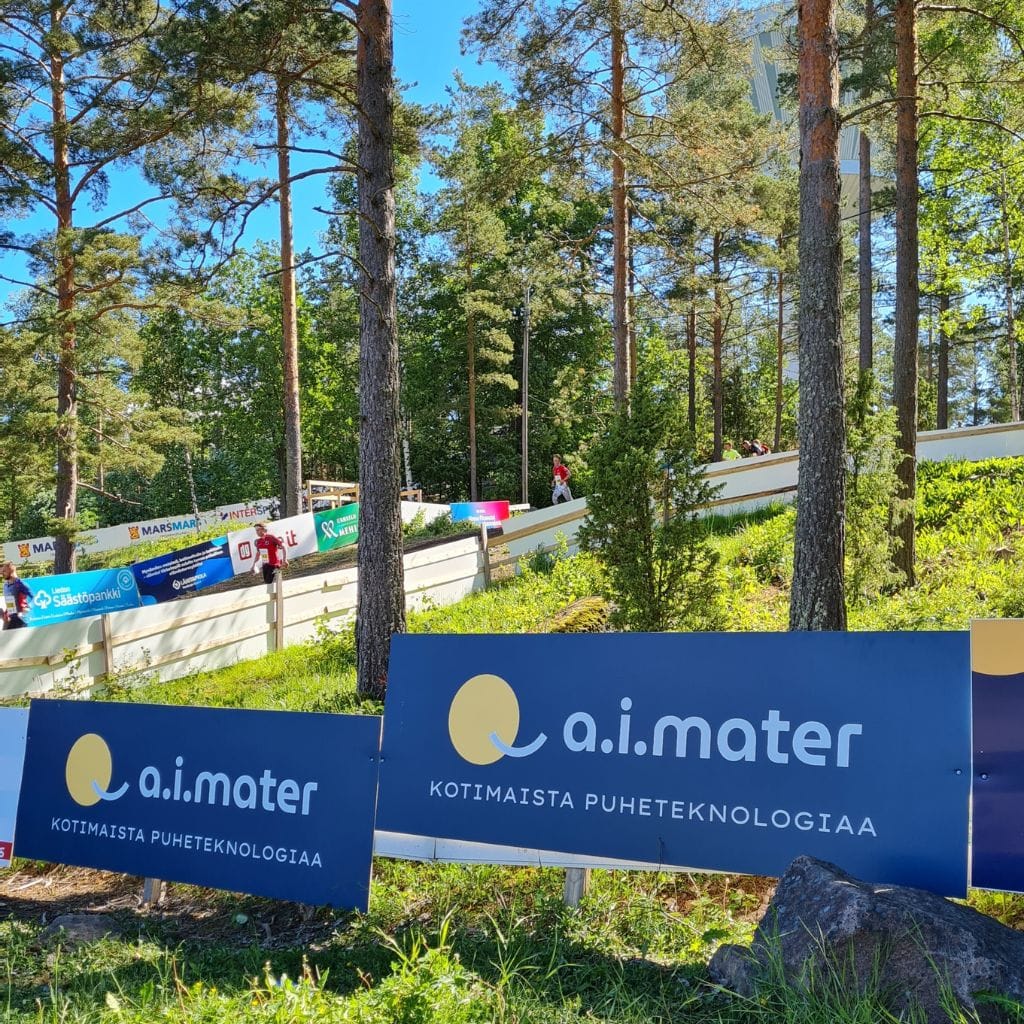 Aimaterin kaksi aitamainosta. Takana kisailijoita juoksemassa maaliin.