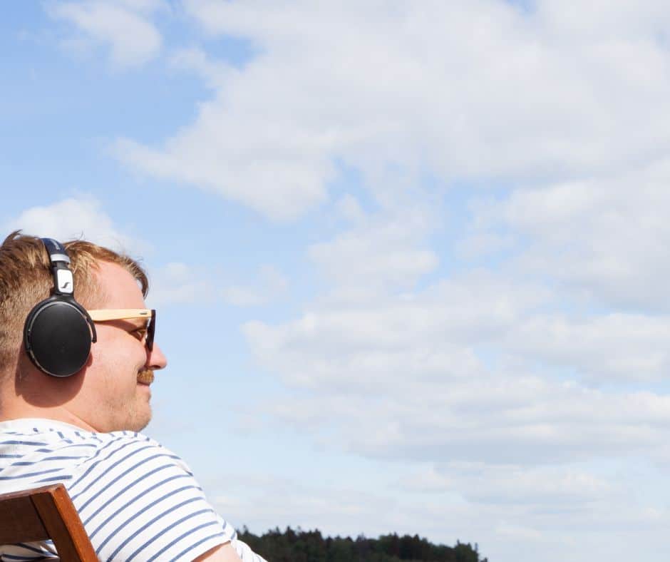 Mies istuu kuulokkeet päässä hymyillen ja katsoo horisonttiin. Taustalla on paljon sinistä taivasta ja harsopilviä.