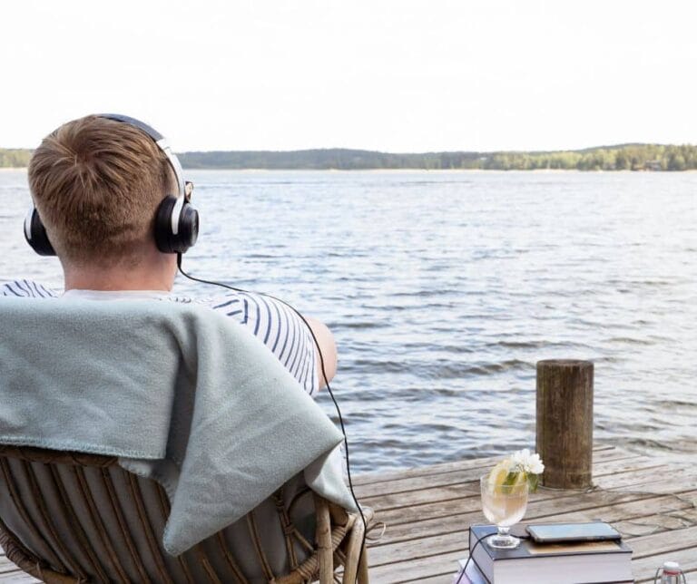 Mies istuu selkä kuvaajaa päin laiturilla korituolissa kuulokkeet päässä. Vieressä on pino kirjoja ja niiden päällä älypuhelin , josta lähtee piuha kuulokkeisiin.
