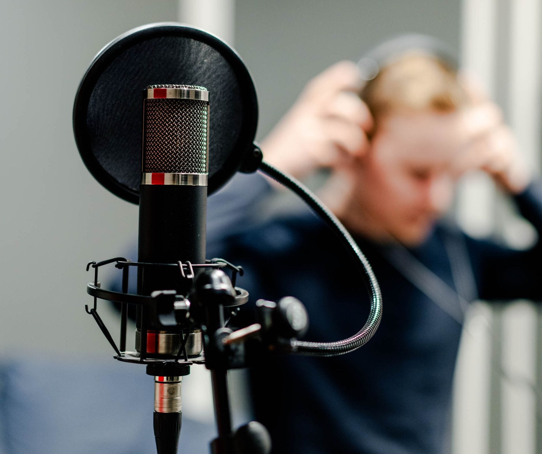 Edessä tarkennettuna studiomikrofoni. Taustalla sumennettuna mies. joka asettaa kuulokkeita päähänsä.